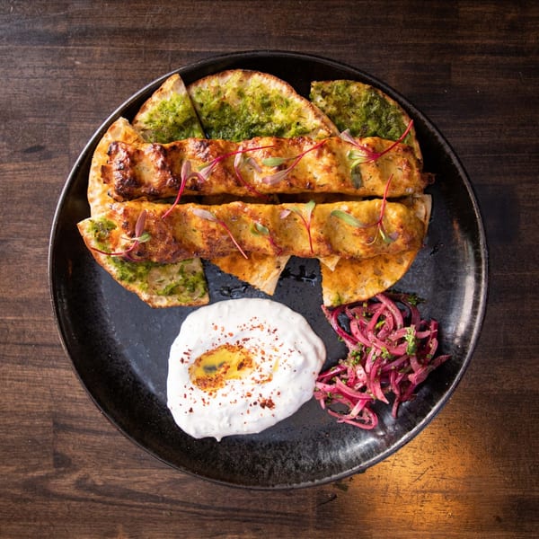 Two chicken kebabs on flatbread with green sauce, a side of yogurt dip, and pickled red onions.