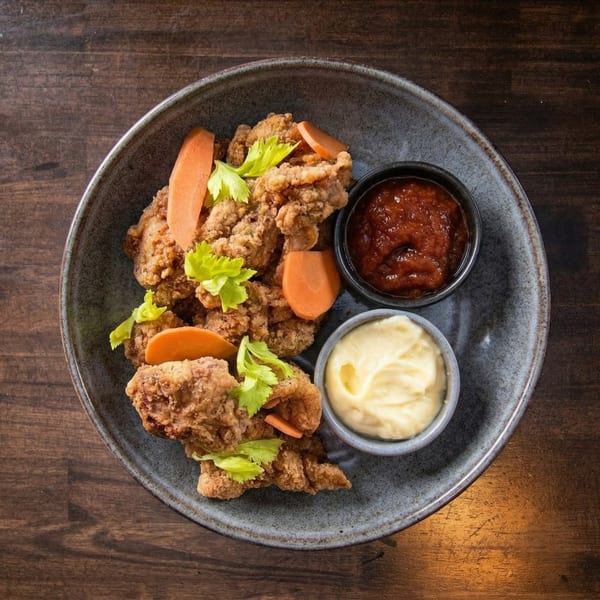 Crispy fried chicken pieces with carrot slices, celery, and two dipping sauces in a gray bowl.