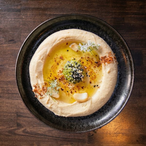 Creamy hummus in a black bowl, drizzled with olive oil, pesto, sesame seeds, and garlic.