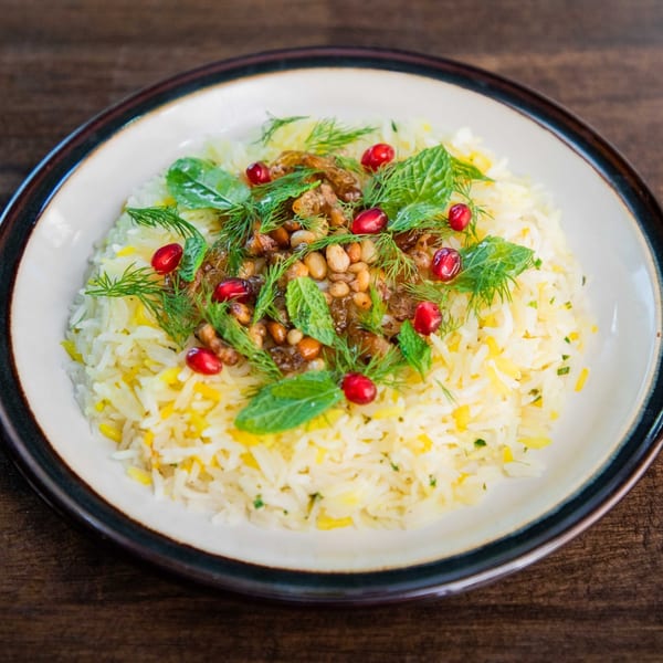 Fluffy white rice topped with pine nuts, pomegranate seeds, mint, and dill on a white plate.